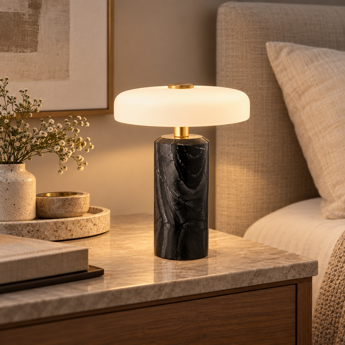 A modern table lamp with a glowing white rounded dome shade and a black marble base with gold accents, styled on a marble bedside table next to a bed.
