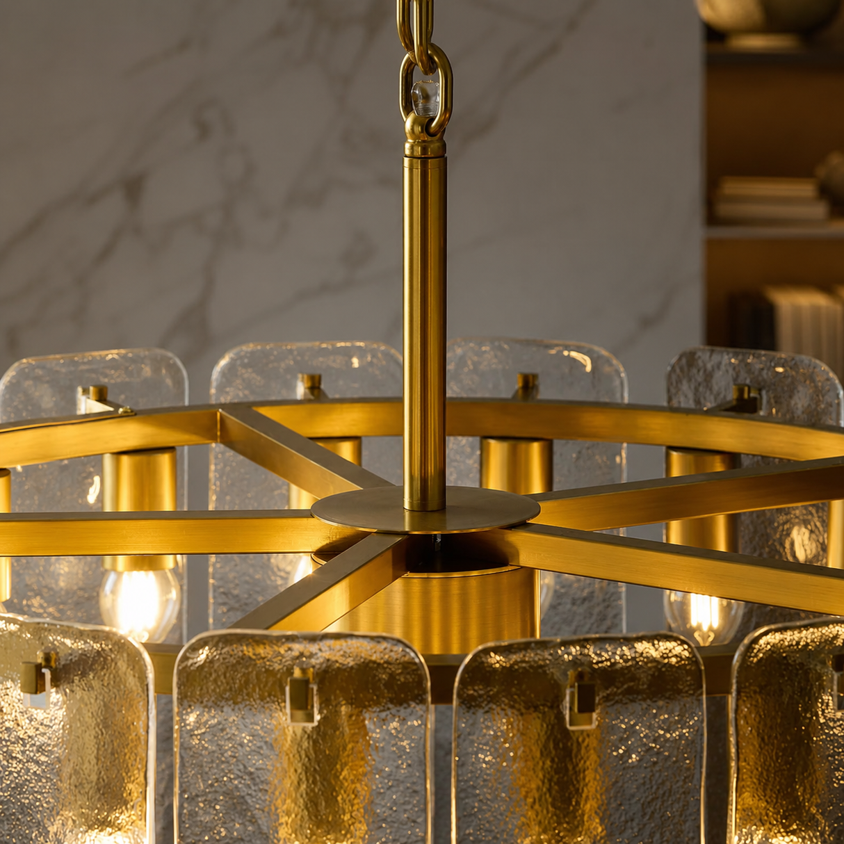 A close-up of a modern chandelier with a brushed gold metal frame, textured clear glass shades, and visible warm light bulbs, shown hanging in an indoor setting.