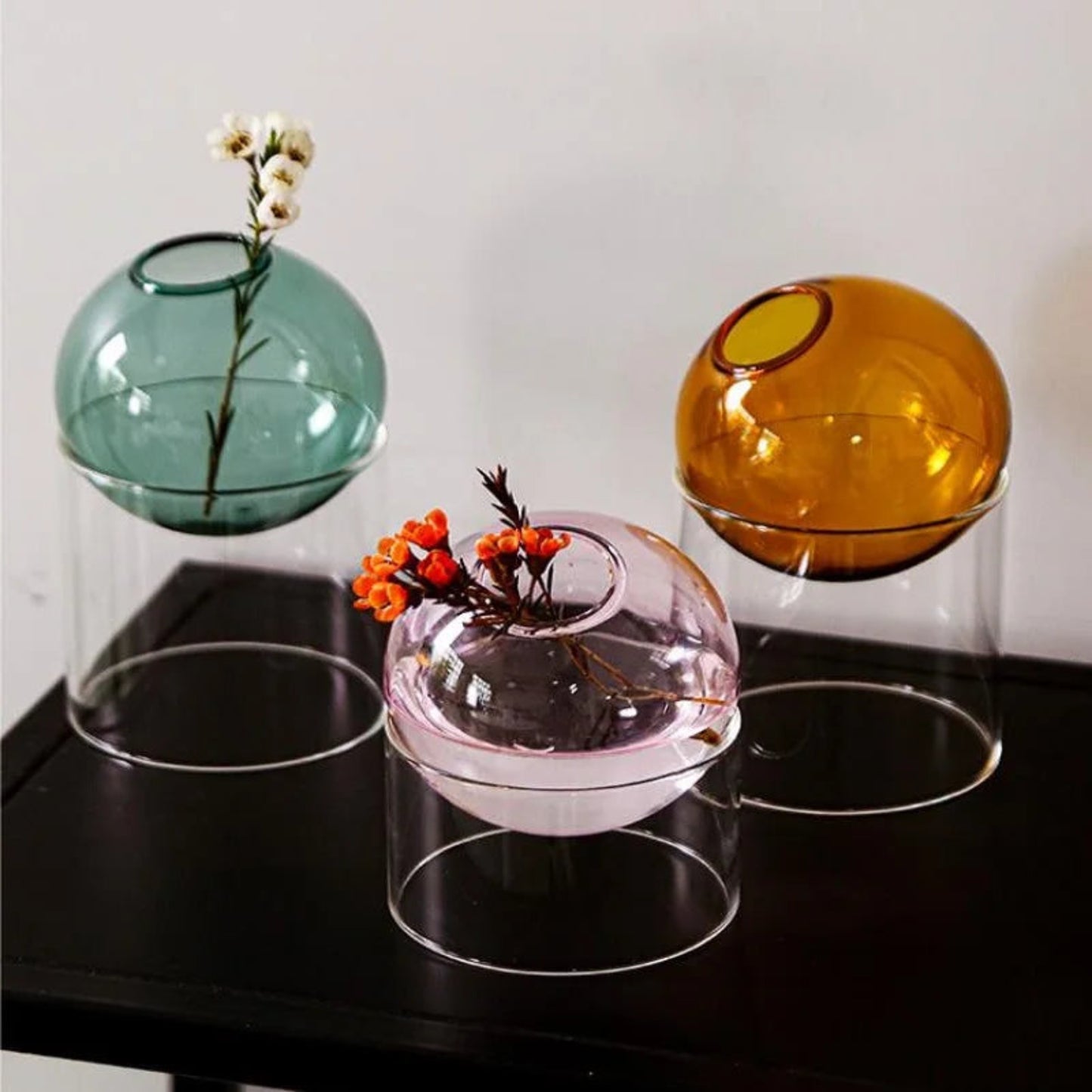 Three transparent glass vases in different colors with floral arrangements on a black surface.