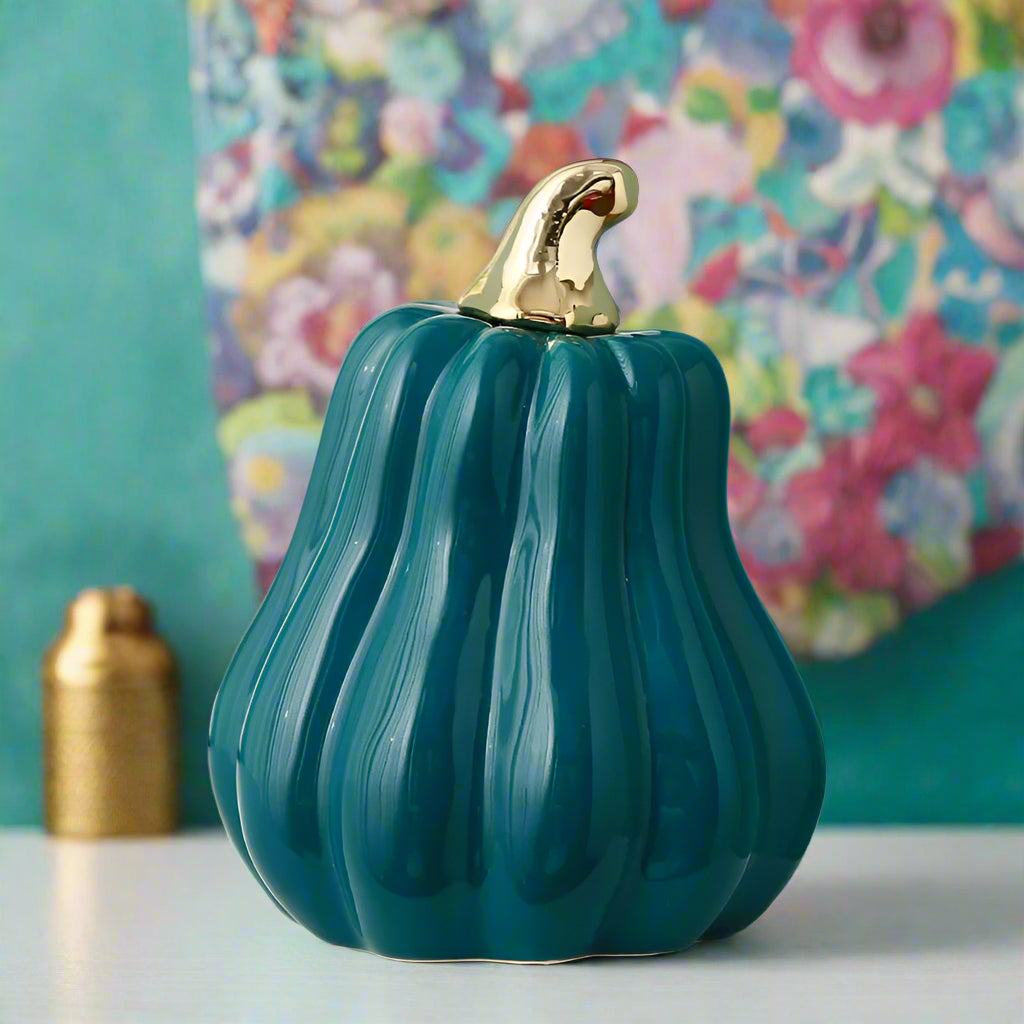 Teal ceramic pumpkin with gold stem on a white surface against a light blue wall.