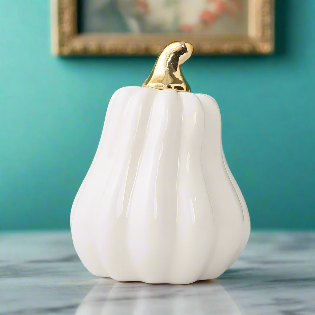 White ceramic pumpkin with gold top on a light surface with flowers in the background
