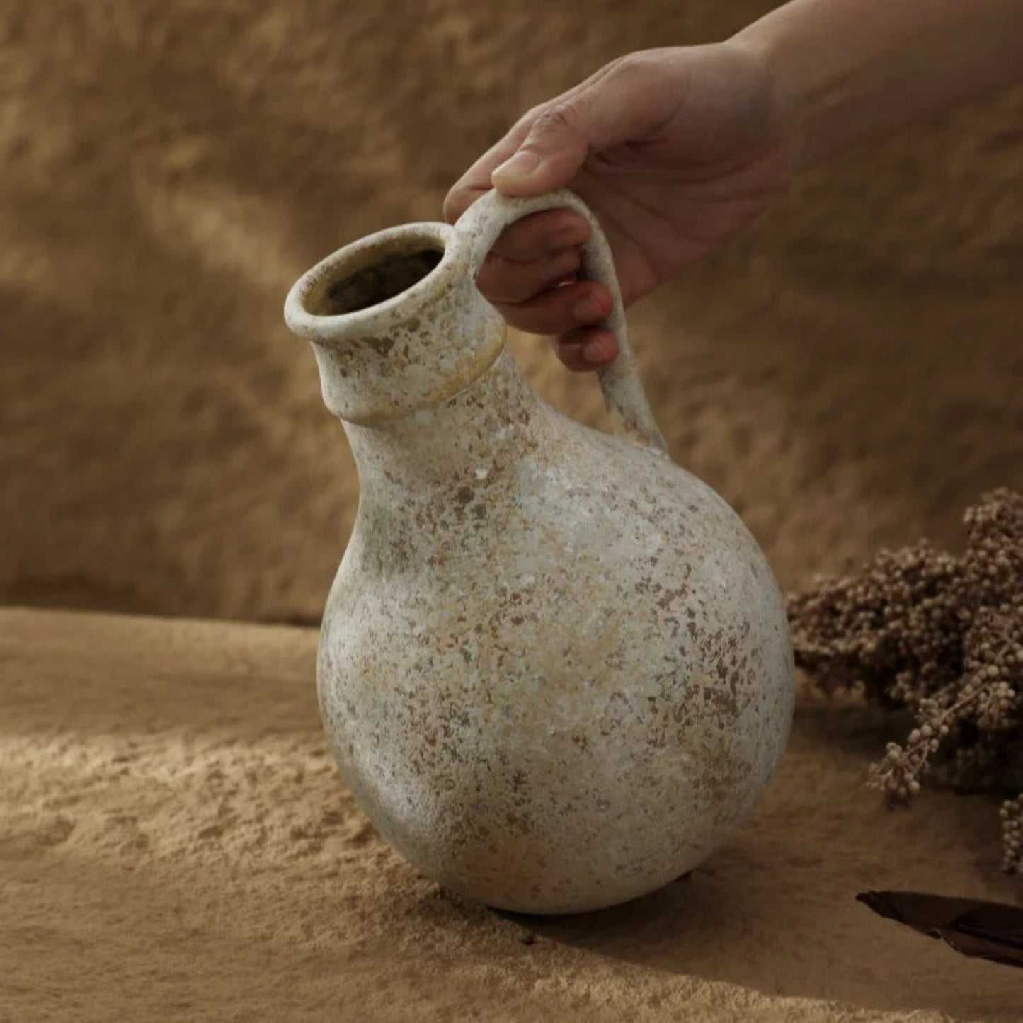 Hand holding a textured ceramic vase on a sandy surface with dried flowers.