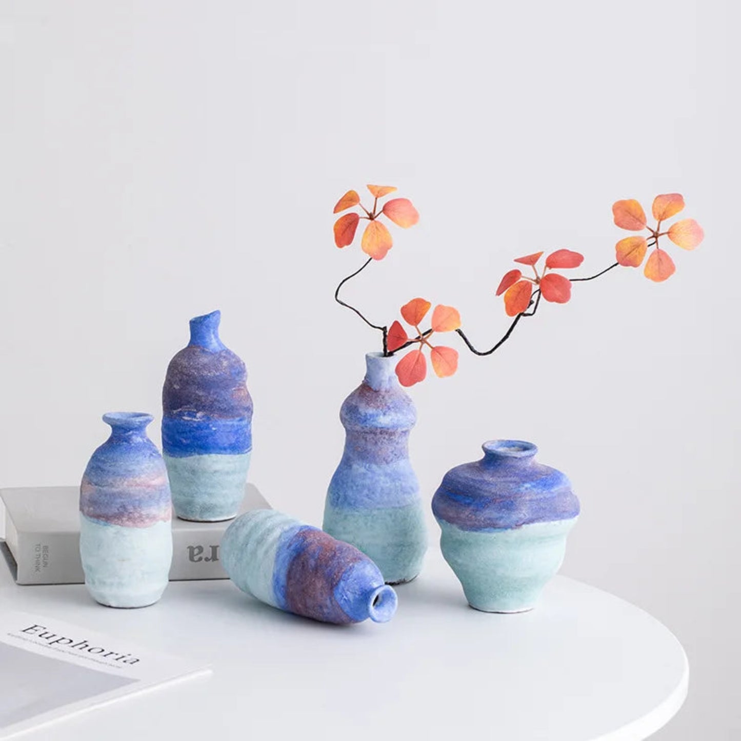 Set of five blue and purple gradient vases on a white surface with a branch of orange leaves.