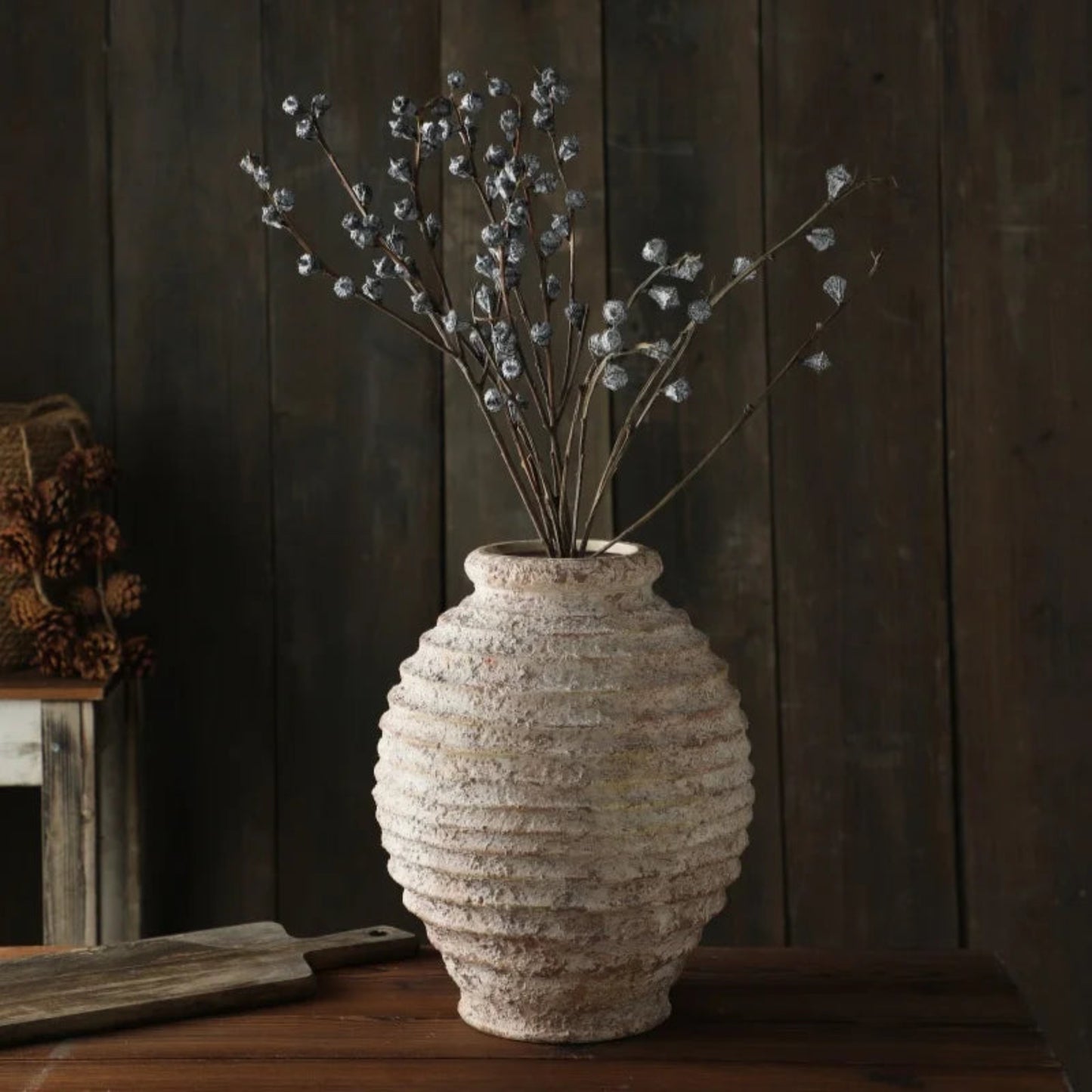 Textured vase with dried branches on a wooden surface against a dark wooden wall.