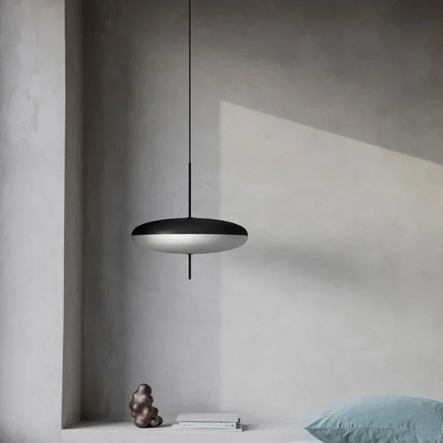 Modern black pendant light hanging in a minimalistic room with concrete walls and a blue pillow.