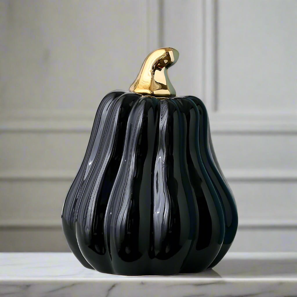 Black decorative pumpkin with a gold stem on a white surface, with green leaves in the background.