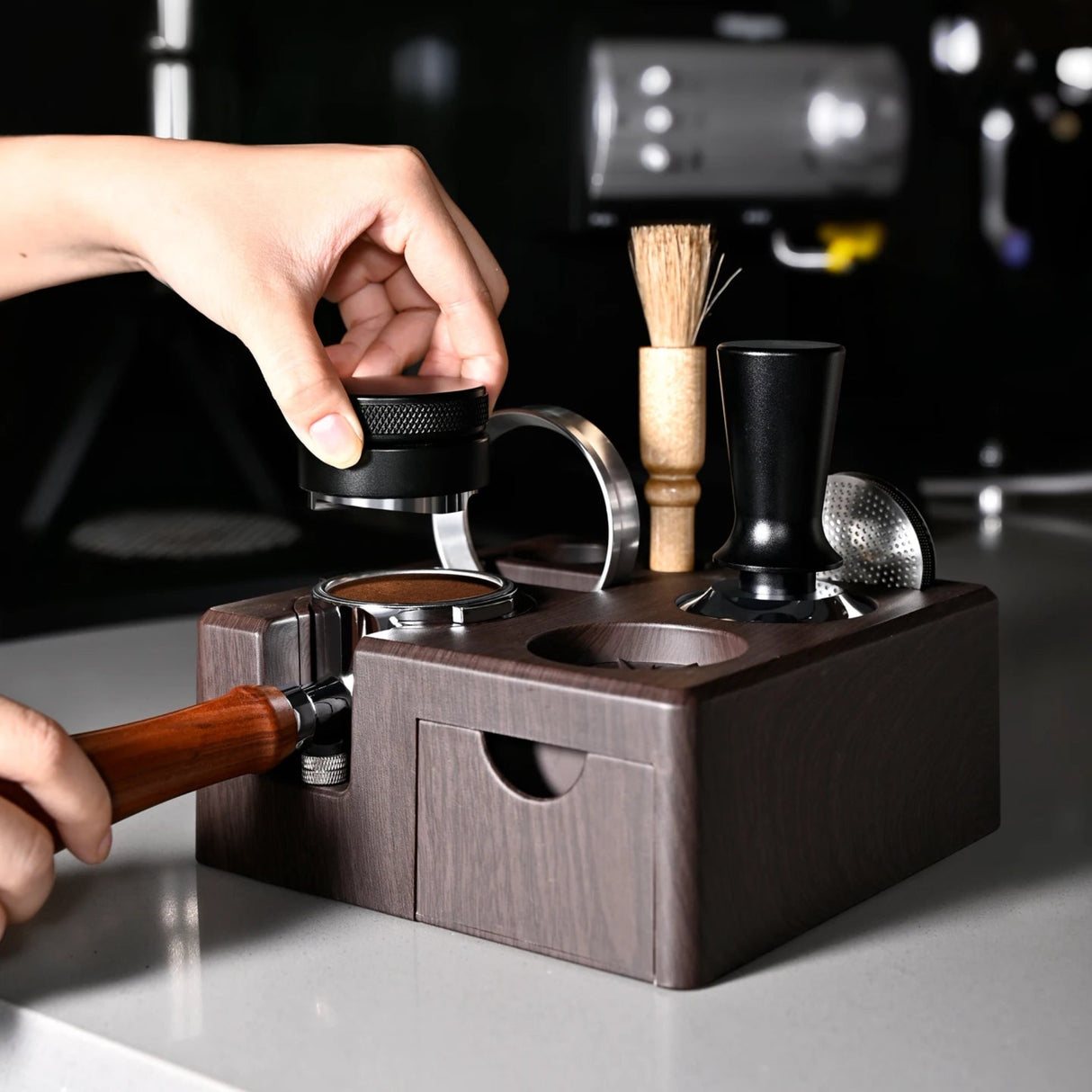 Hand using a wooden tool to operate a coffee grinder on a dark surface.