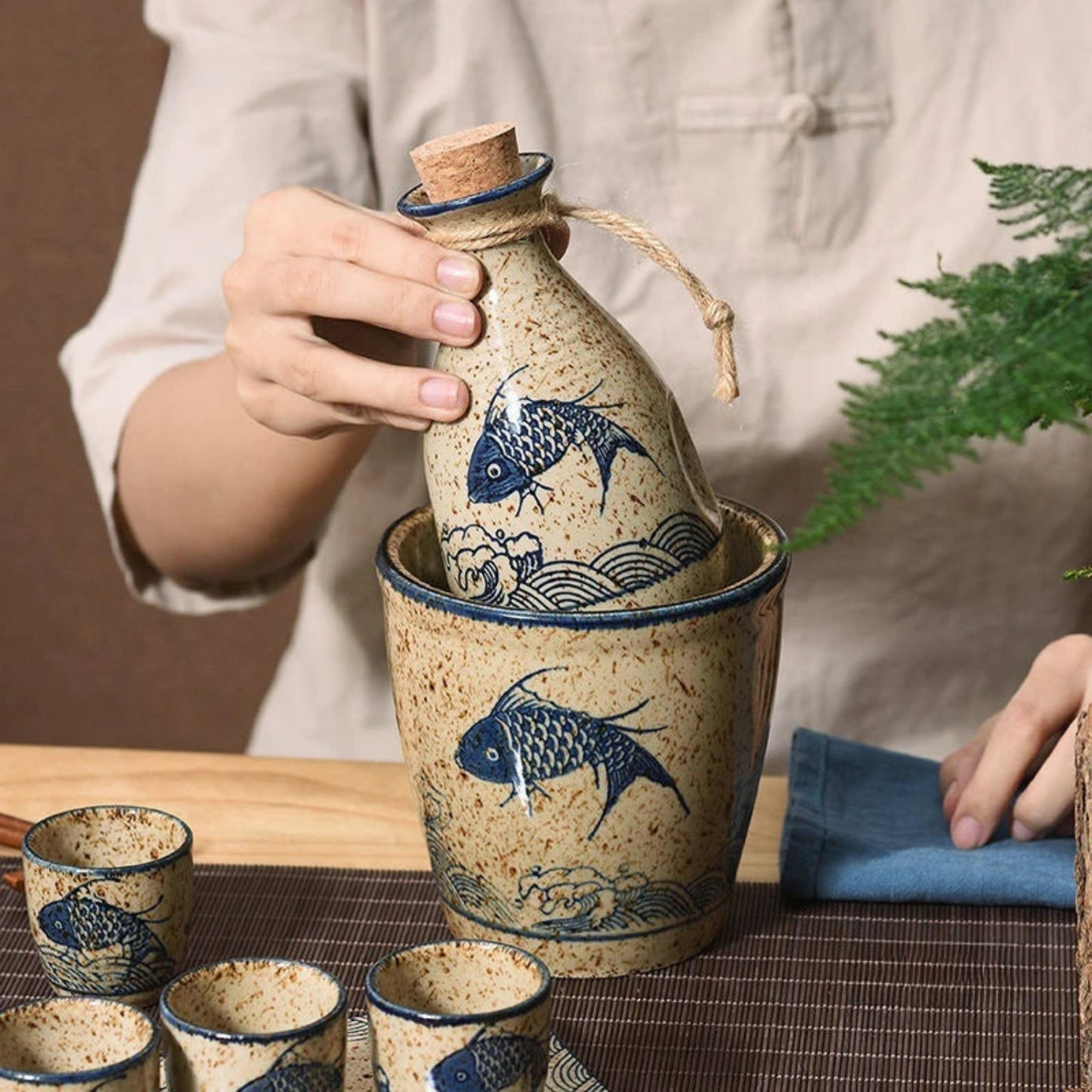 Ceramic bottle with cork stopper and blue fish design, held by a person with a plant in the background.