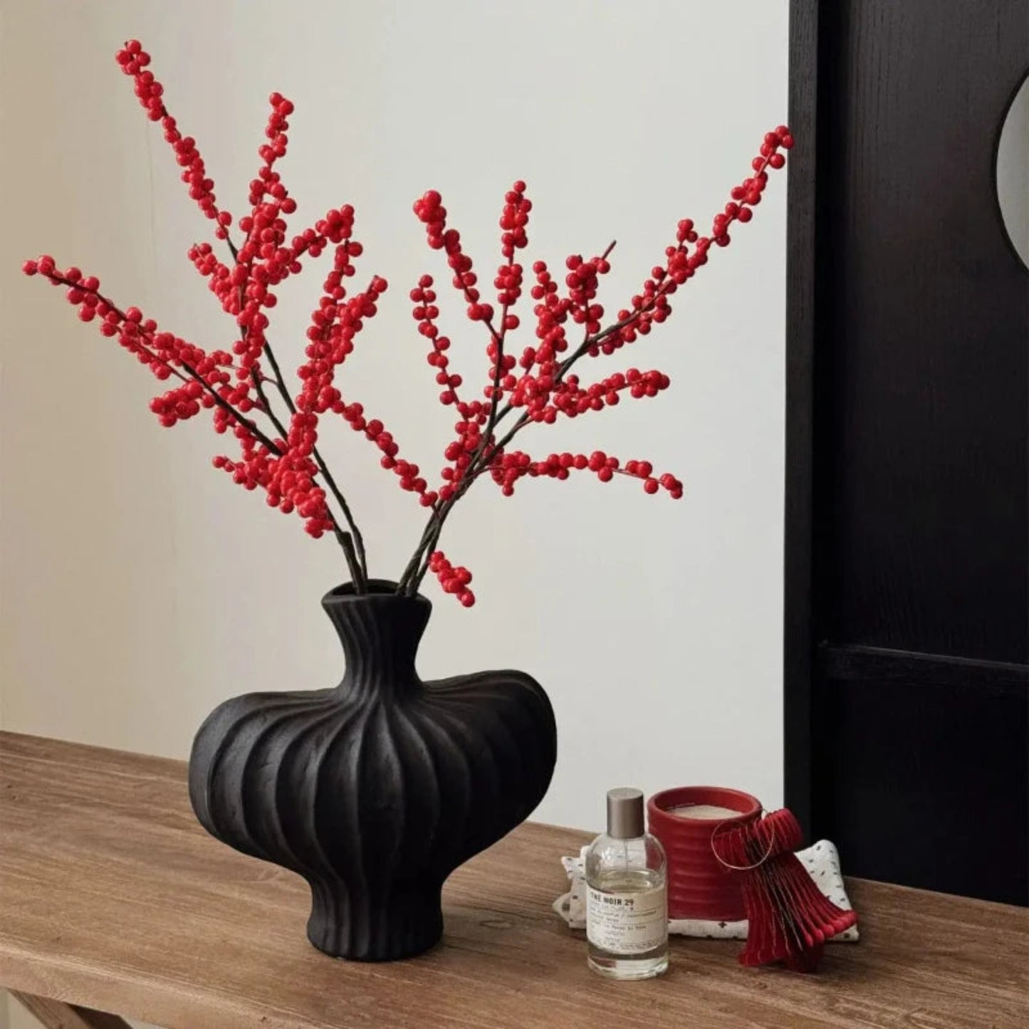 Black vase with red branches on a wooden surface next to a candle and small bottle.