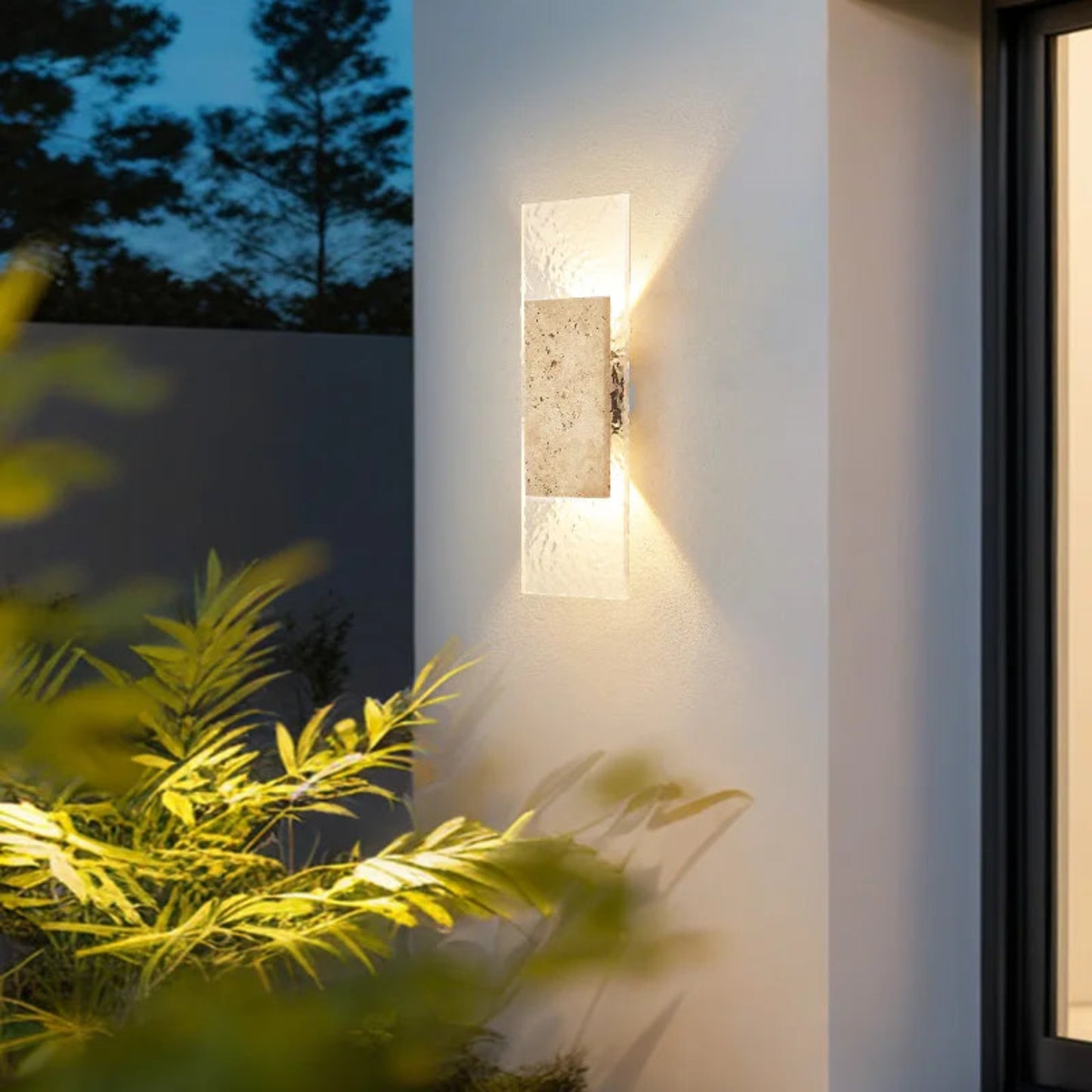 Modern outdoor wall light fixture on a building exterior with plants in the foreground.