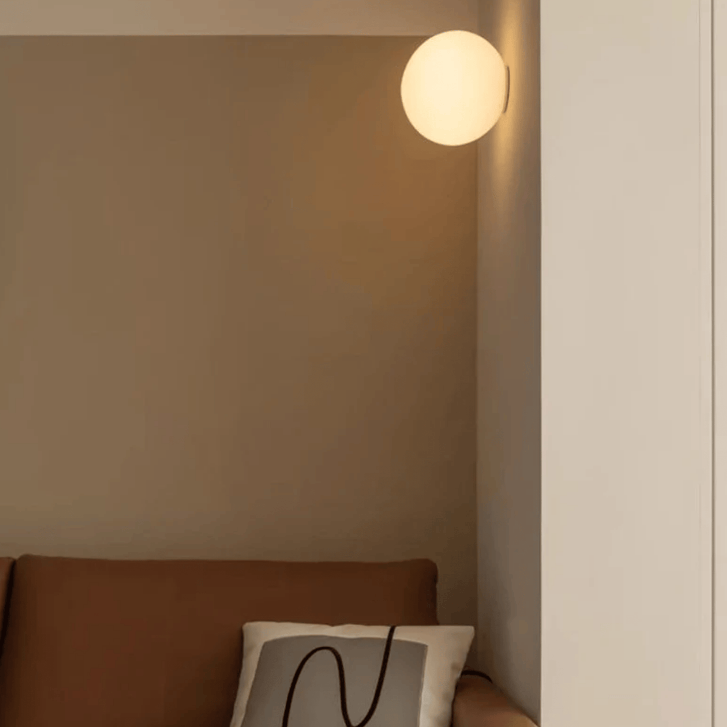 Wall-mounted light fixture casting a warm glow on a neutral wall with a brown sofa and white pillow.