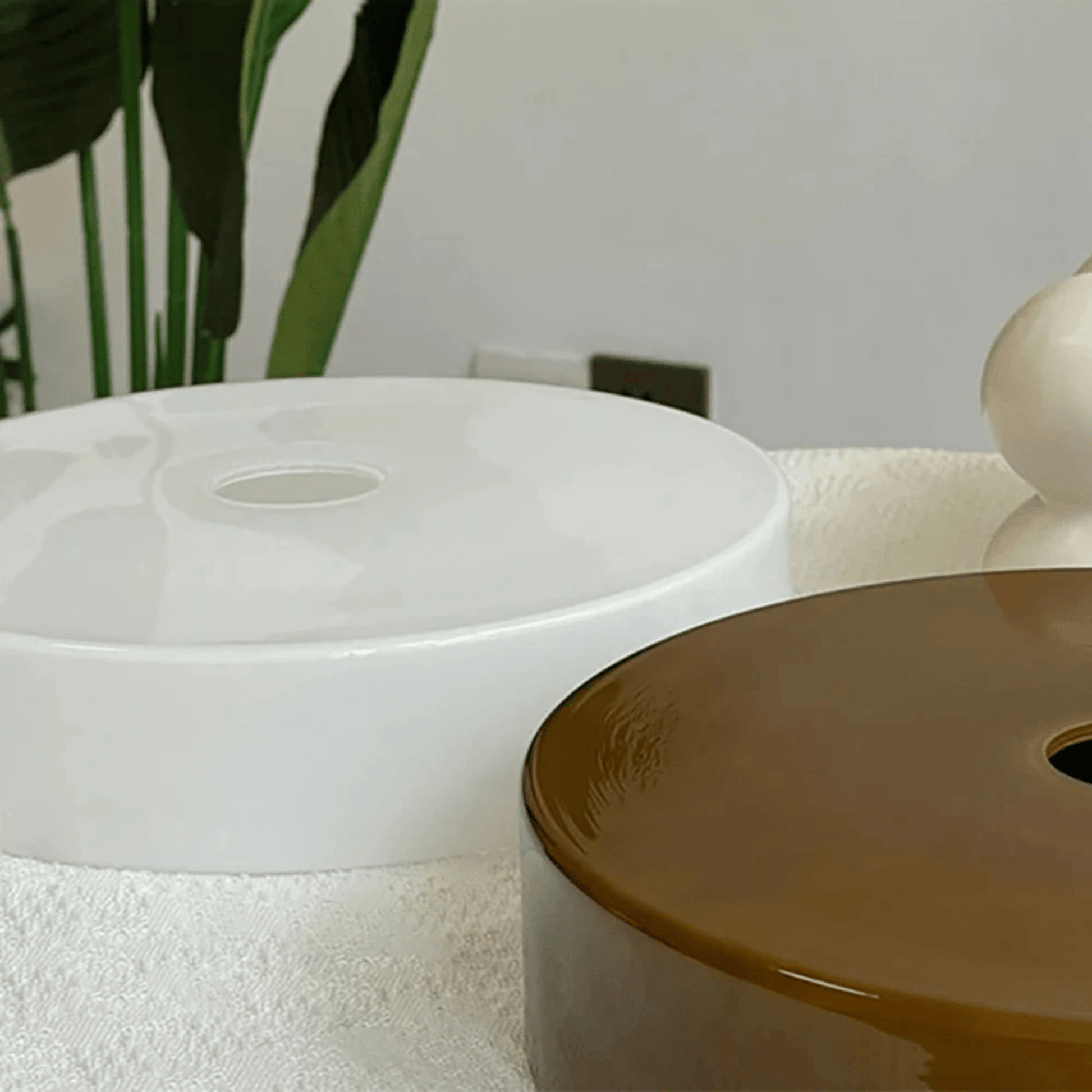 Two ceramic lamp shade one white and one brown, on a textured surface with a plant in the background.