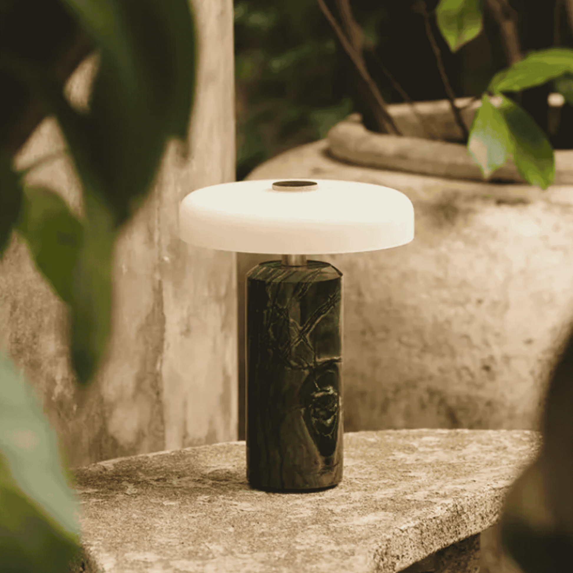 Table lamp with a wooden base and white lampshade on a stone surface with greenery in the background