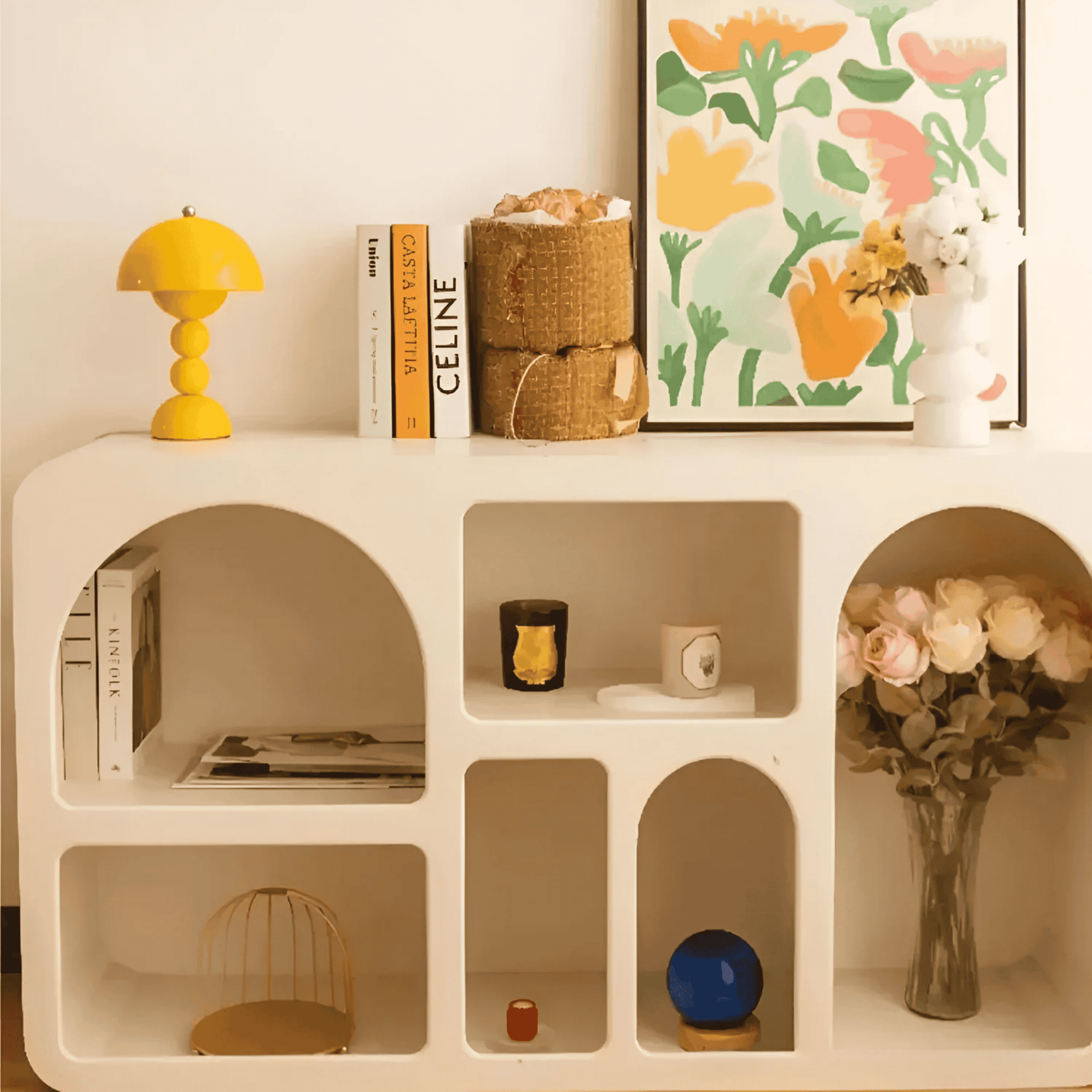 White shelving unit with decorative items including a yellow lamp, books, a candle, and a vase with flowers.