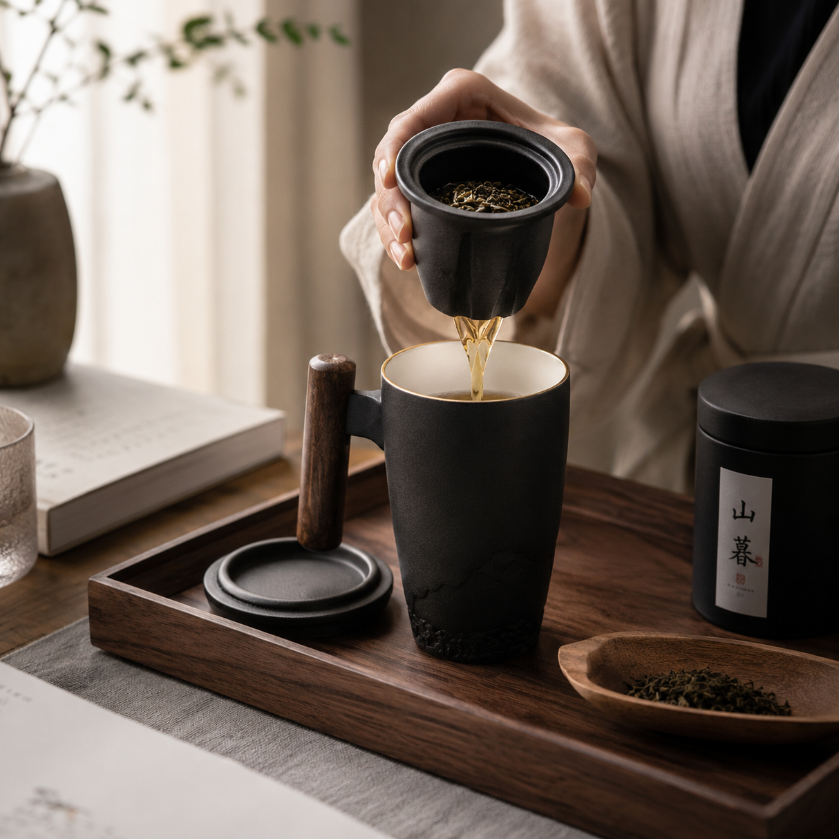 A person in a beige robe pours tea from a black filter holder into a black ceramic mug with a wooden handle, set on a wooden tray with a tea tin and a small dish of loose leaves.