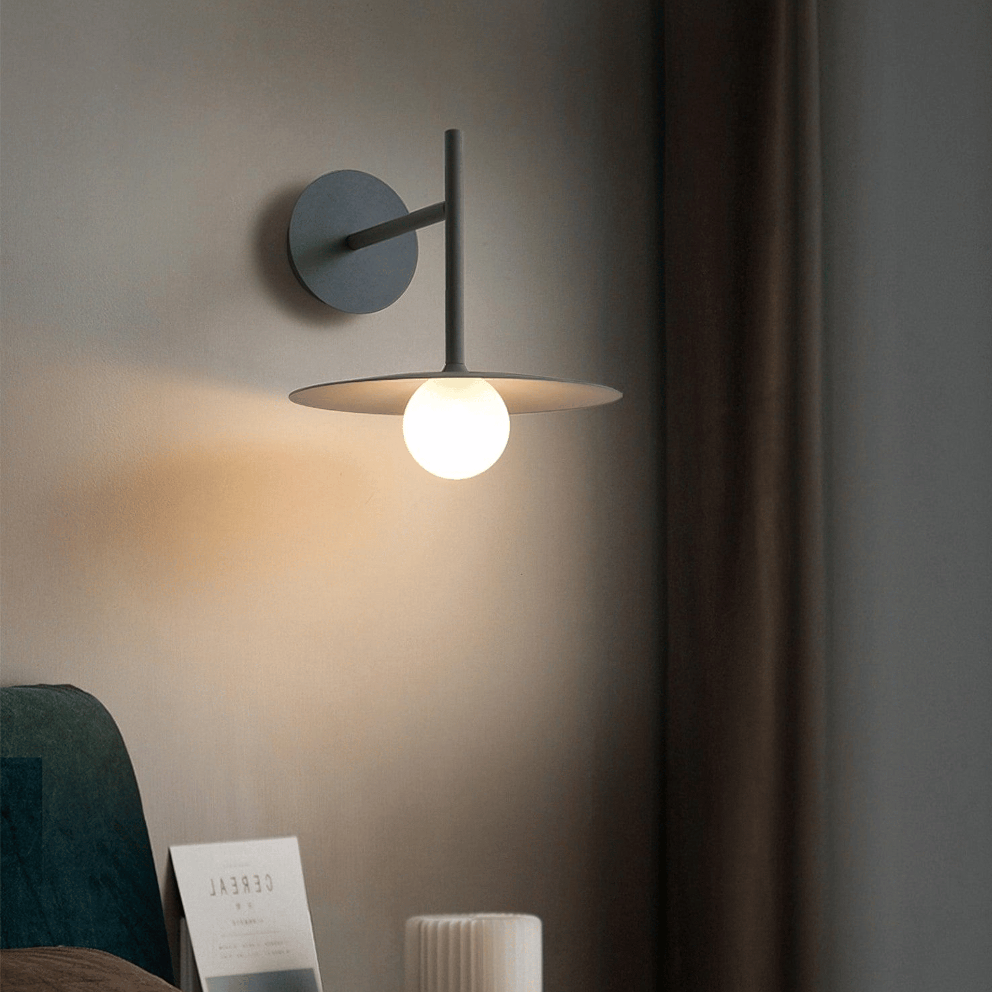 Modern wall-mounted lamp on a neutral wall with a blurred chair and magazine in the foreground.