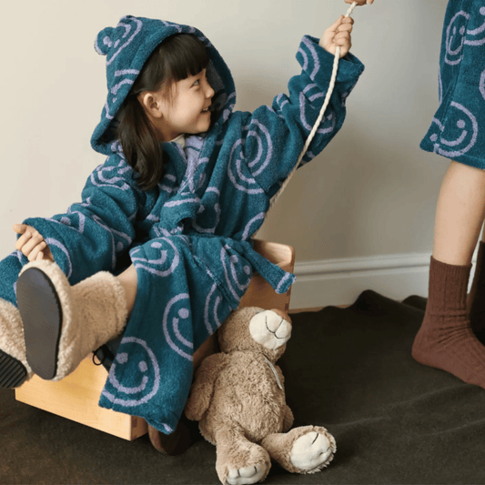 Child in a blue robe with white patterns sitting on a wooden horse toy with a teddy bear.