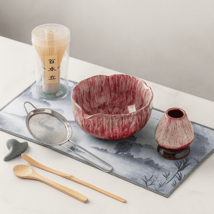 Red ceramic bowl with matching small pitcher on a decorative mat with tea-making tools.