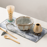 Tea-making set with ceramic bowl, whisk, spoon, and small vase on a decorative mat.