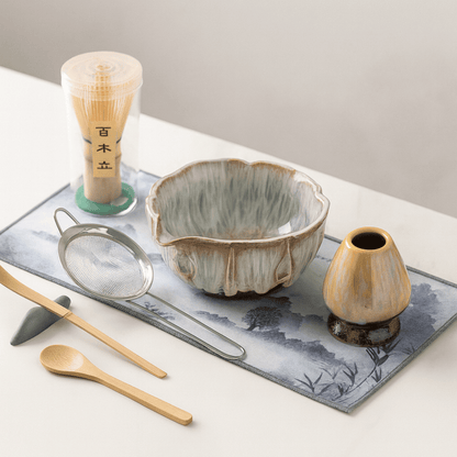 Tea-making set with ceramic bowl, whisk, spoon, and small vase on a decorative mat.