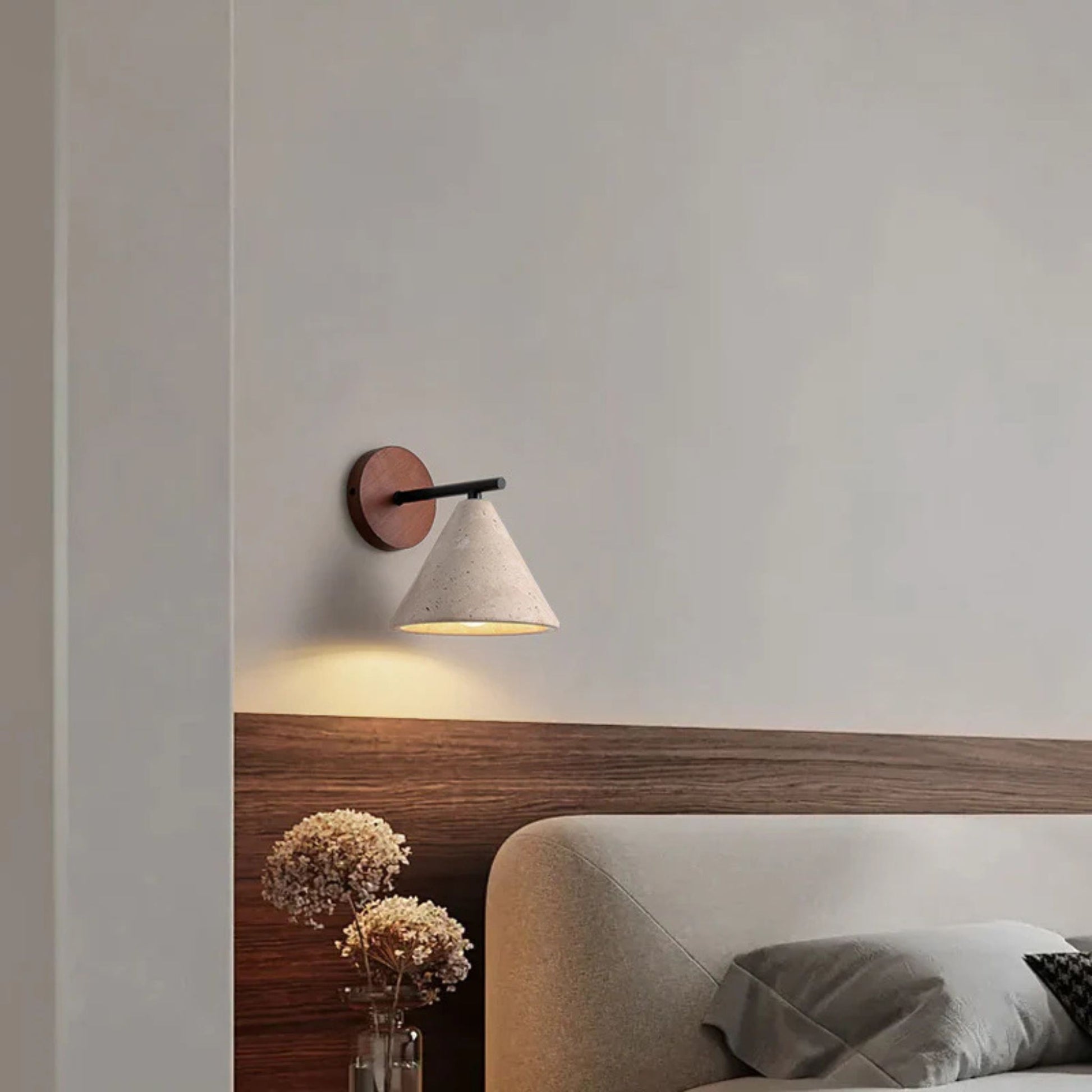 A modern wall sconce with a wooden base and white cone-shaped shade, glowing warmly above a bed in a minimalist bedroom.