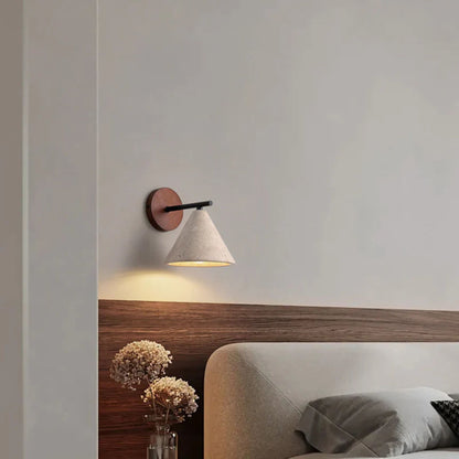 A modern wall sconce with a wooden base and white cone-shaped shade, glowing warmly above a bed in a minimalist bedroom.