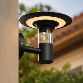 Black outdoor wall light fixture on a building exterior with blurred greenery in the background.
