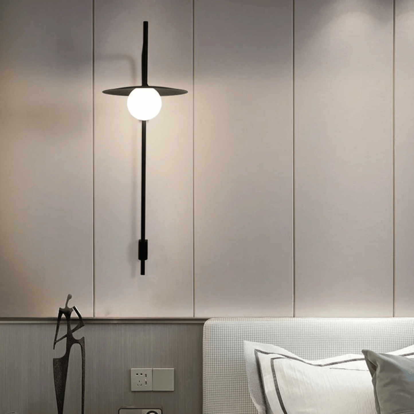 Modern wall-mounted light fixture on a neutral wall with a bed in the foreground.