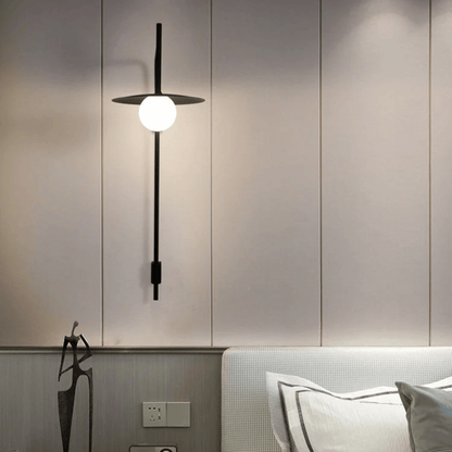 Modern wall-mounted light fixture on a neutral wall with a bed in the foreground.