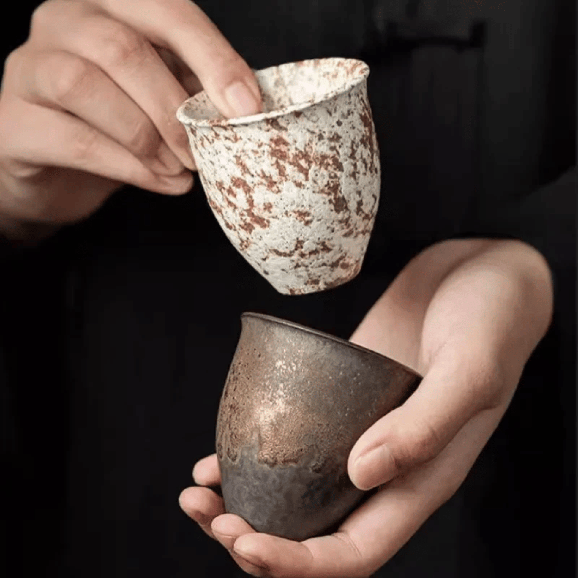 Two hands holding ceramic cups with a speckled glaze against a black background