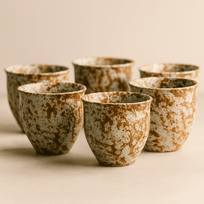 Set of six ceramic cups with brown and white speckled glaze on a beige background