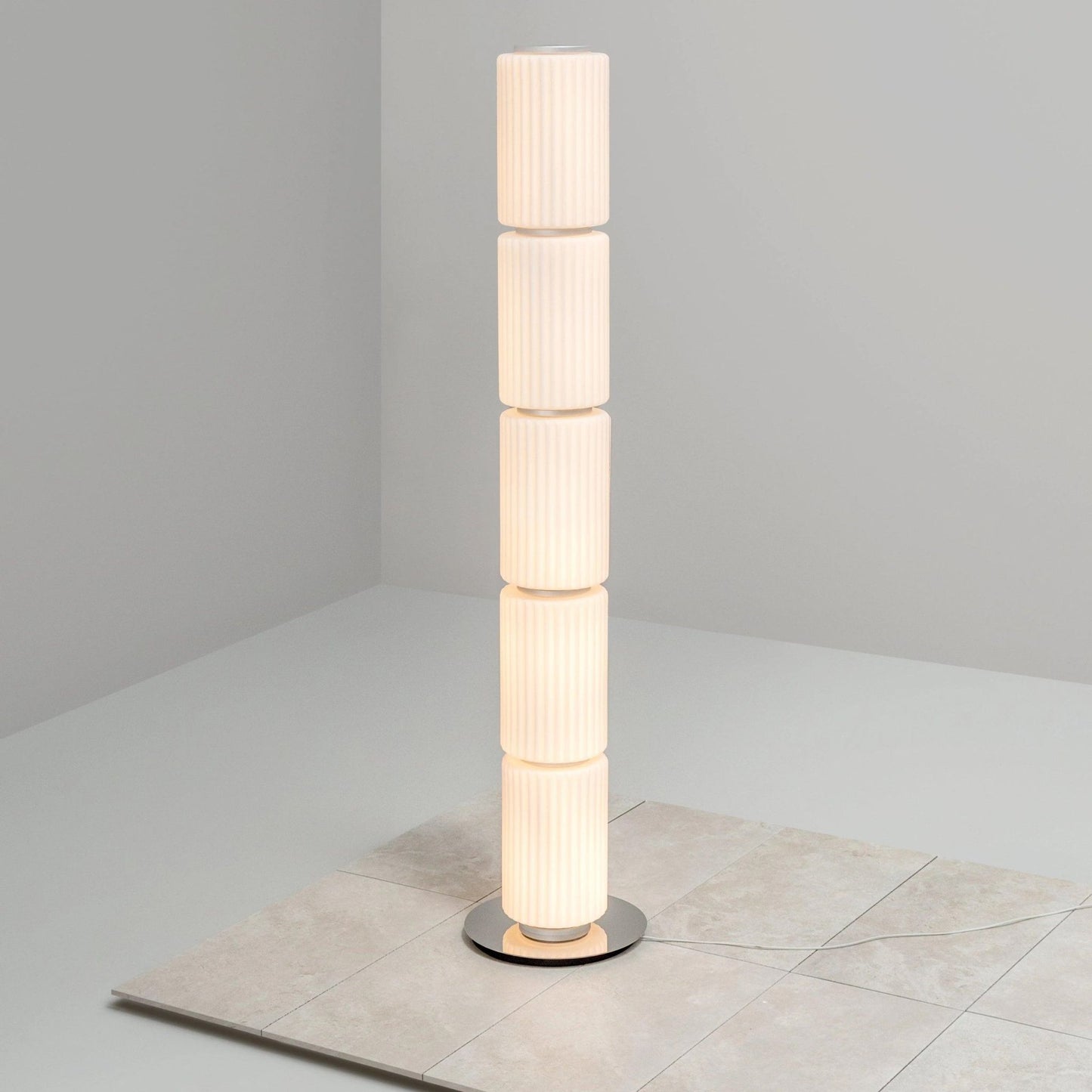 Vertical multi-segment ribbed-glass floor lamp illuminated on a smooth white floor.