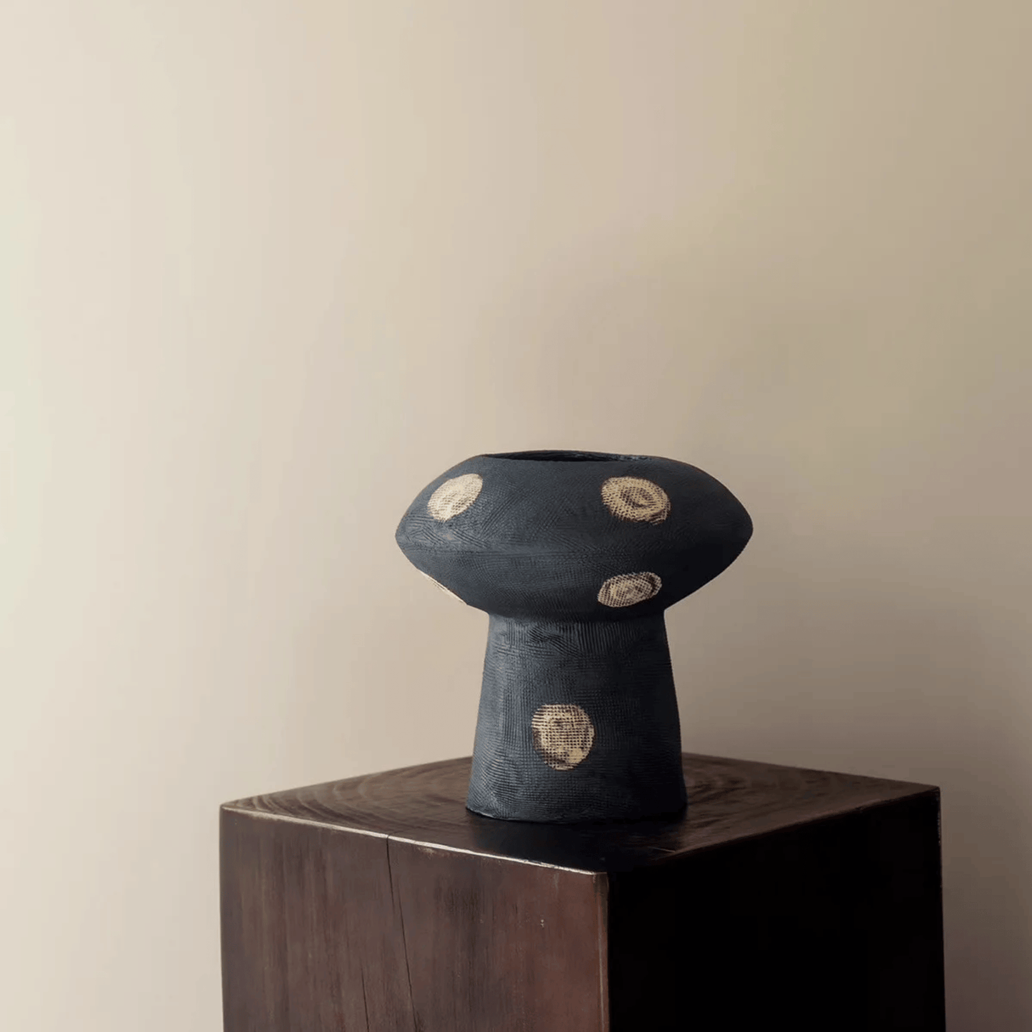 Black ceramic vase with gold circular patterns on a wooden pedestal against a beige wall