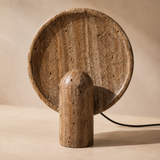 A sculptural table lamp made from natural travertine stone, featuring a round backplate and a cylindrical pedestal base, shown on a neutral beige background.