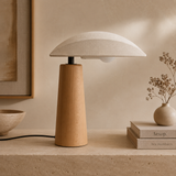 A modern mushroom-shaped table lamp with a textured off-white dome shade and a tapered light wood base, styled on a stone surface next to books and a small vase of dried flowers.