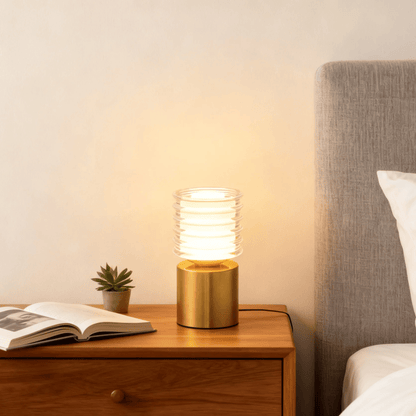 Gold table lamp on a wooden nightstand next to an open book and small plant, with a bed in the background.