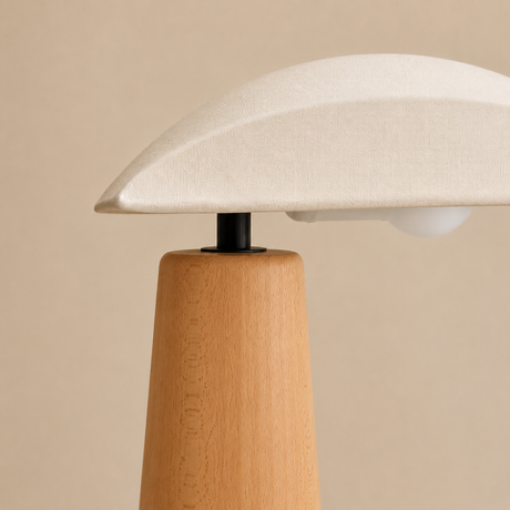A close-up of a table lamp with a light beige fabric dome shade, a natural wood tapered base, and a black metal connector, set against a solid beige background.