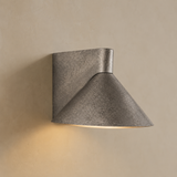 A modern wall-mounted sconce with a textured gray metal finish, featuring a square backplate and a tapered cone shade, casting a warm light against a beige wall.