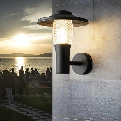 Modern black outdoor wall light with textured glass glowing at sunset near a waterfront