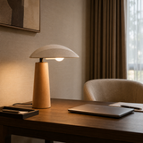 A modern table lamp with a light wood cylindrical base and a white dome-shaped shade sits on a dark wood desk in a warm, softly lit room. A closed laptop, notebook, and chair are visible nearby, with curtains and a window in the background.
