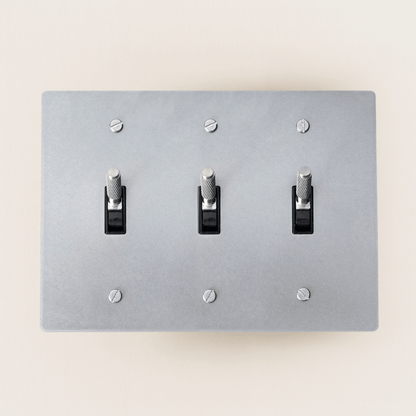 Gray light switch panel with three toggle switches on a beige wall.