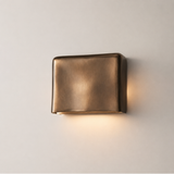 A modern square wall sconce with a brushed bronze finish, mounted on a light beige wall and casting a warm glow downward.