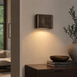 A modern rectangular wall sconce with a dark bronze finish, mounted on a beige wall and casting a warm light downward. The scene includes a dark wood console table with books, a wooden bowl, and a large vase with greenery.