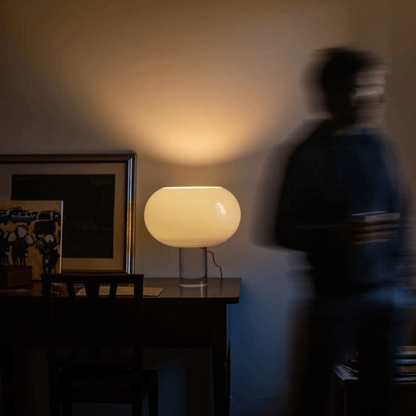 Person standing in a dimly lit room with a table lamp and framed picture.