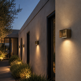 A modern square wall-mounted outdoor light fixture in a brushed brass finish, illuminated and mounted on a stucco house exterior at dusk.
