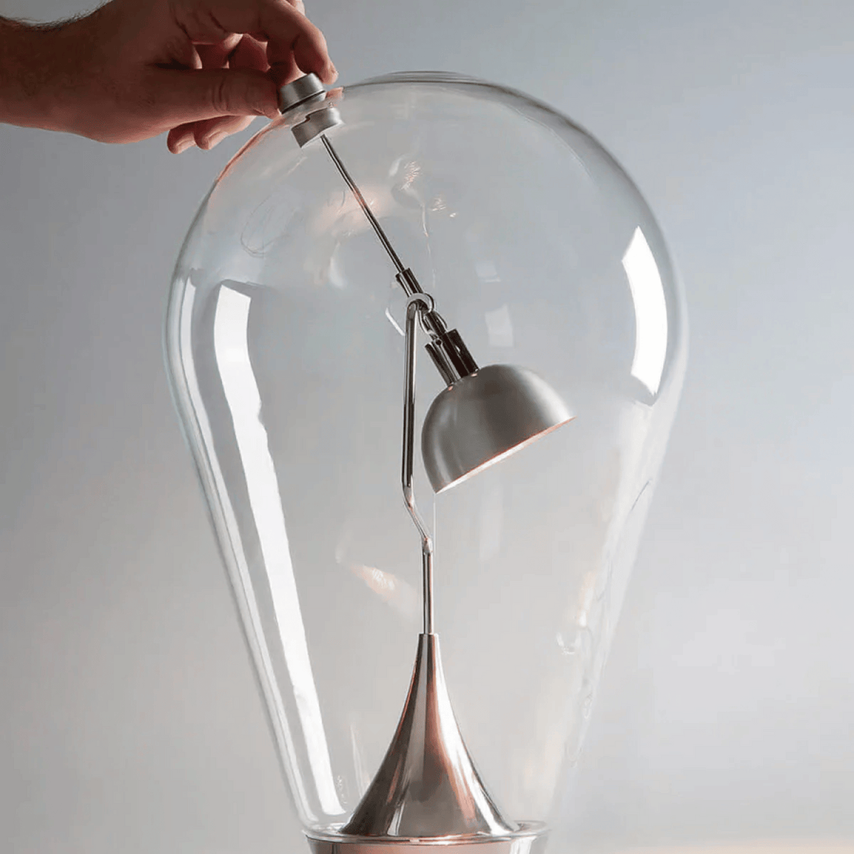 Clear glass light bulb with a hand adjusting a white lampshade against a plain background