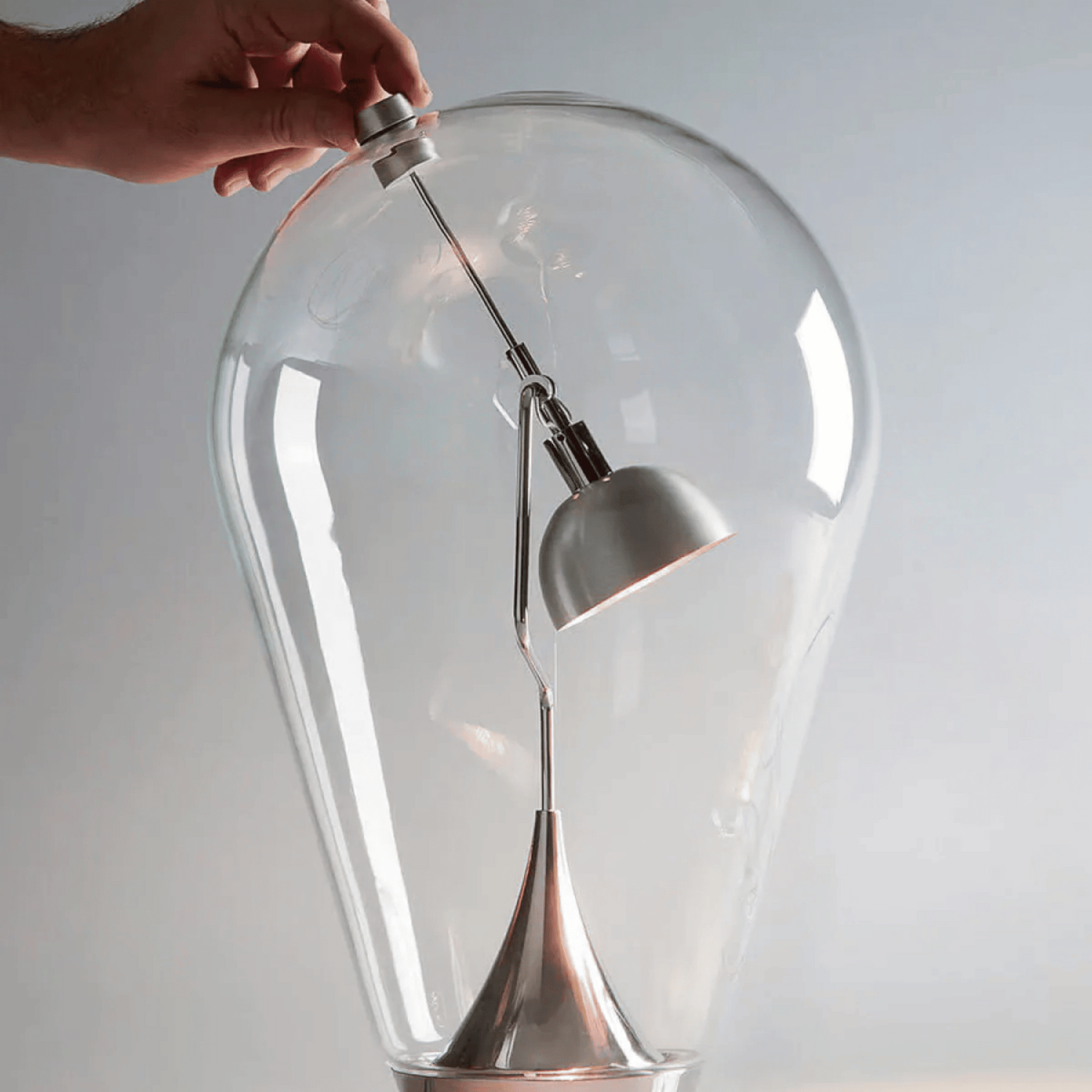Clear glass light bulb with a hand adjusting a white lampshade against a plain background