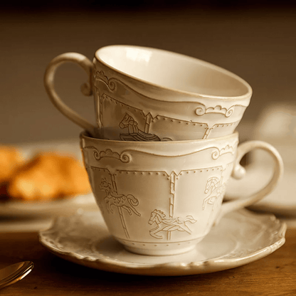 Two stacked ceramic cups with decorative patterns on a saucer, with pastries in the background.