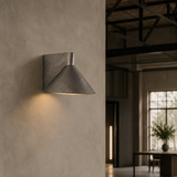 A modern wall-mounted cone-shaped sconce with a textured dark gray finish, mounted on a textured plaster wall and casting a warm light downward, with an industrial loft interior visible in the background.