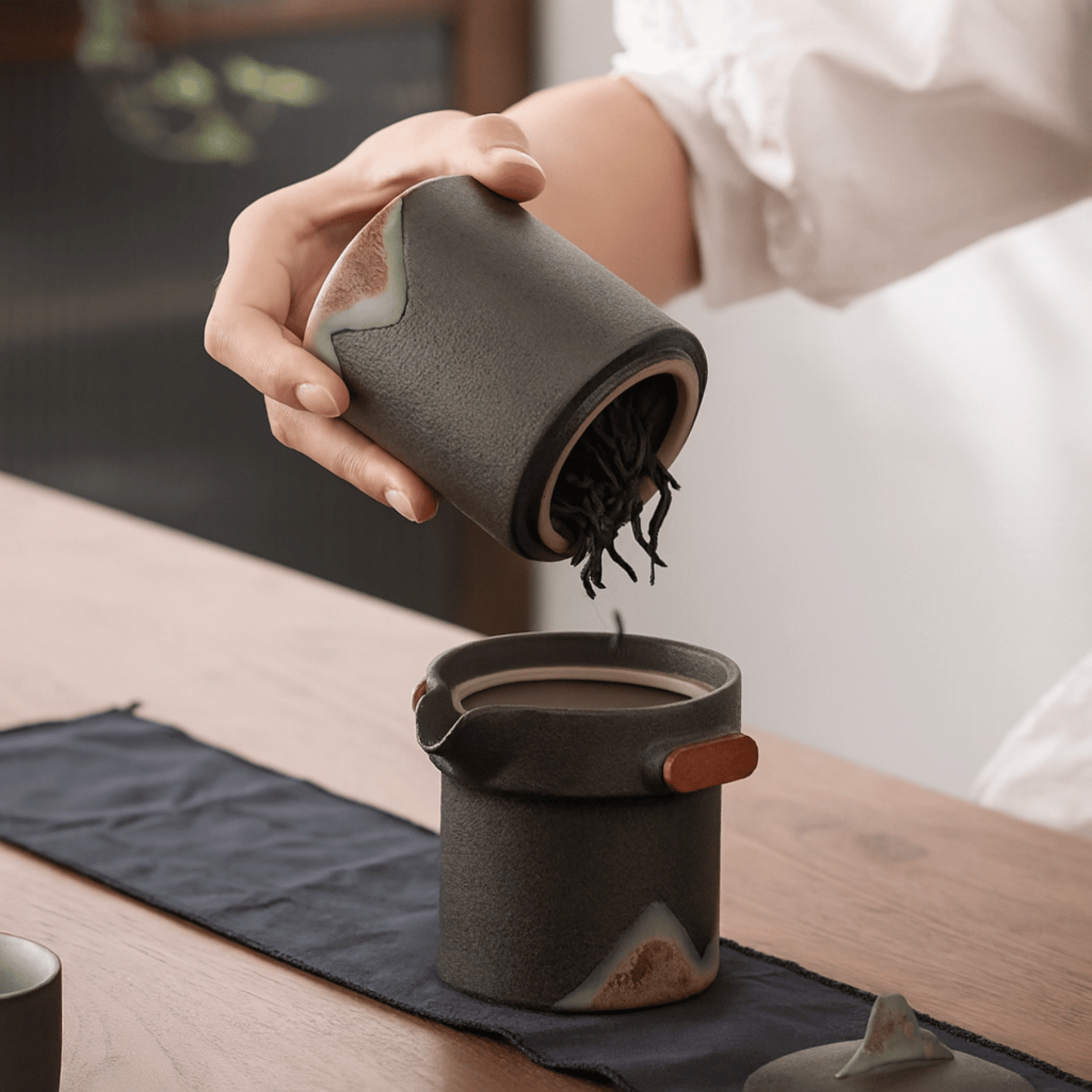 Person pouring from a ceramic container into another on a wooden table.